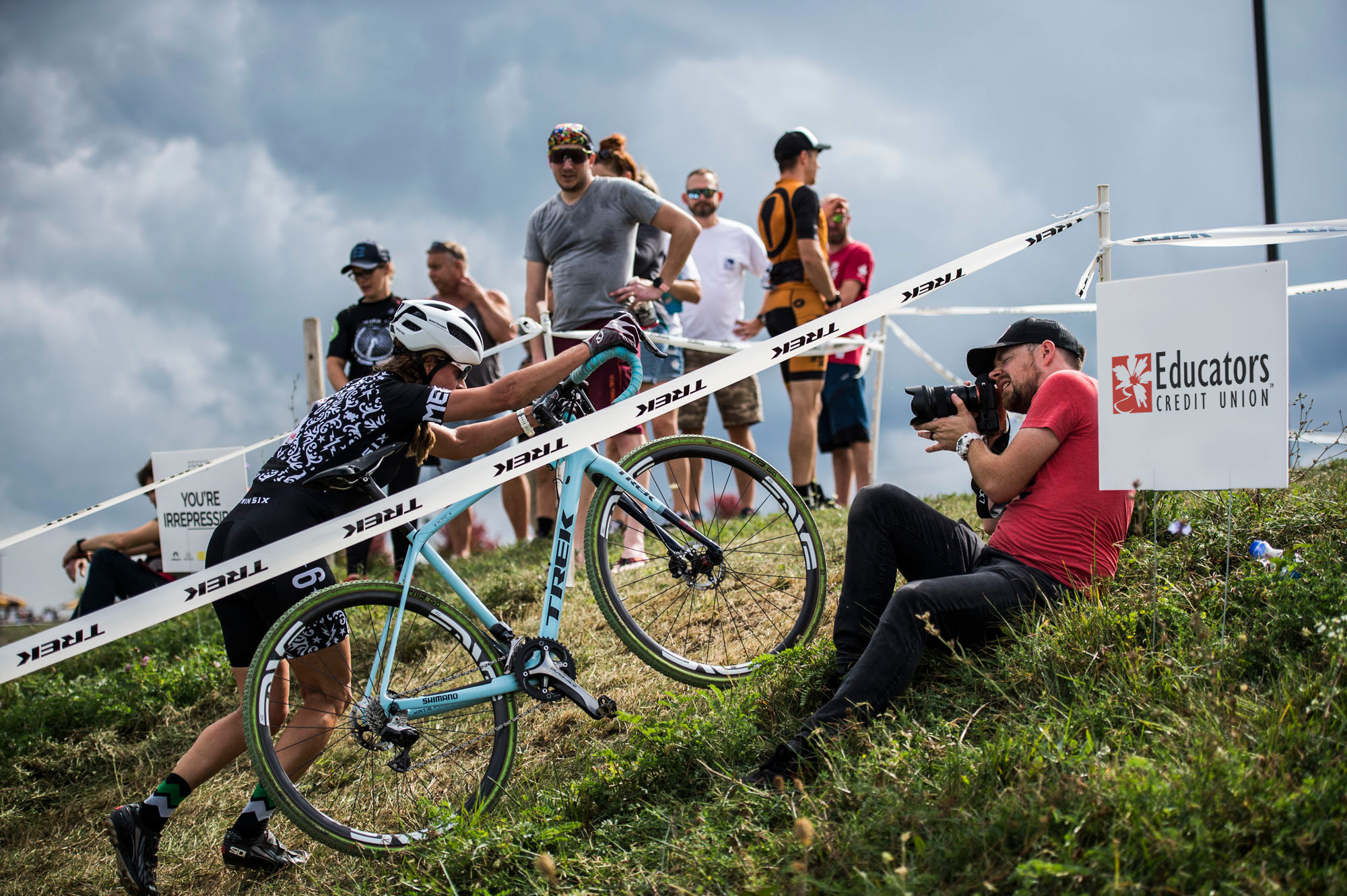 A woman pushes a bike up a very steep gravel hill as a peanut gallery looks on.