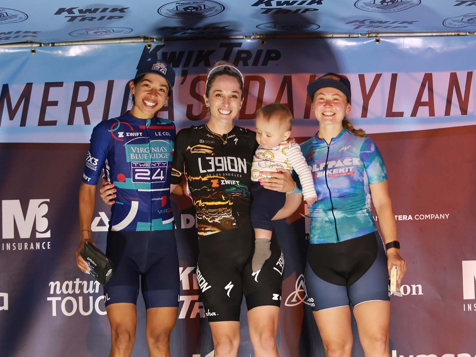 Three bike racers stand on the podium with a baby.