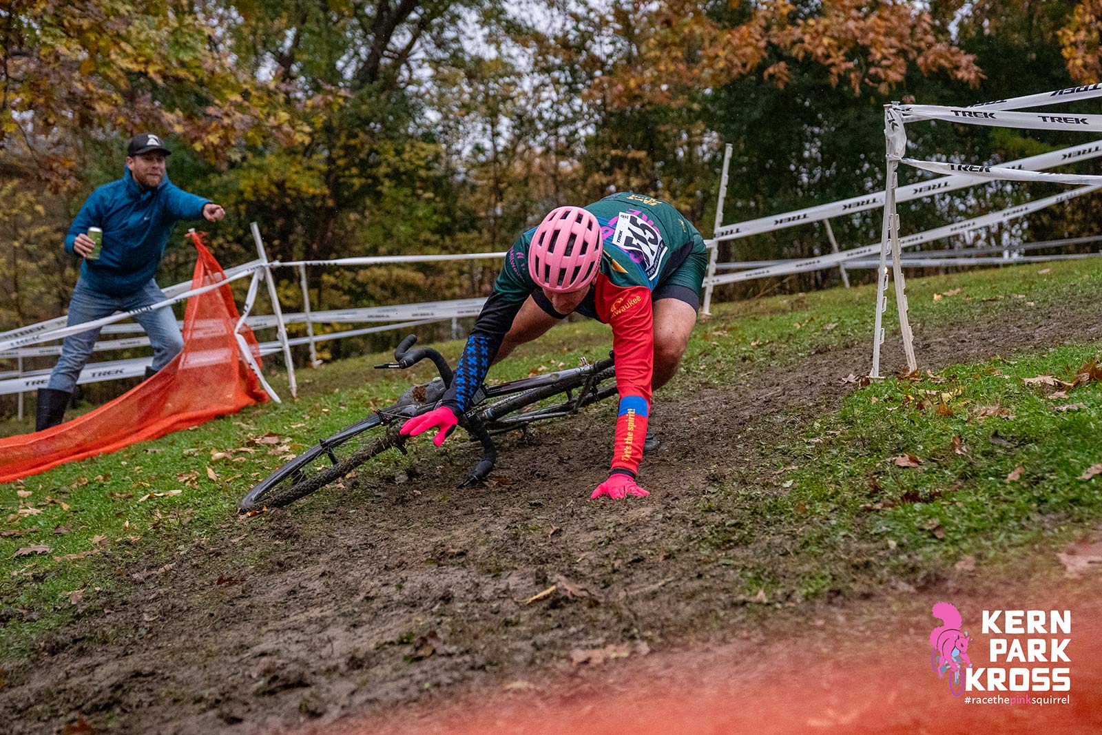 A man falls off his cyclocross bike on a slippery, muddy, off-camber turn, while another points and laughs.