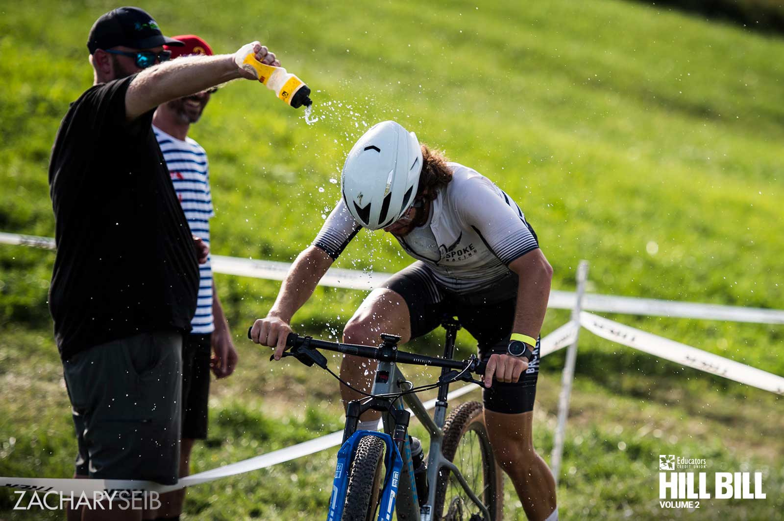 A man squirting a water bottle onto a hot cyclocross racer as they remount their bike.