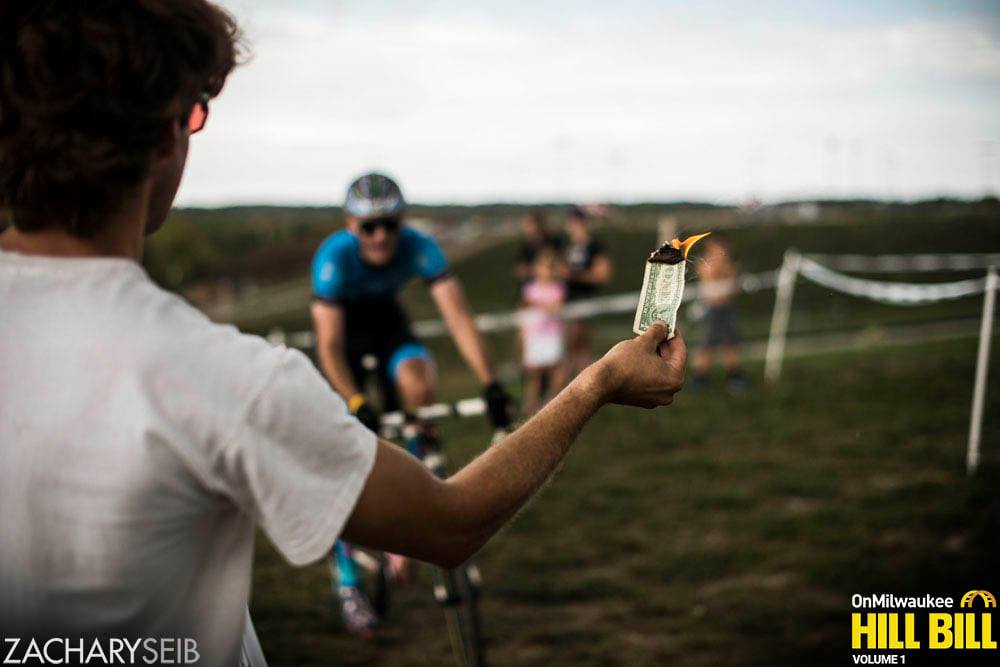 A man dangles a burning dollar bill in front of an approaching cyclocross racer.