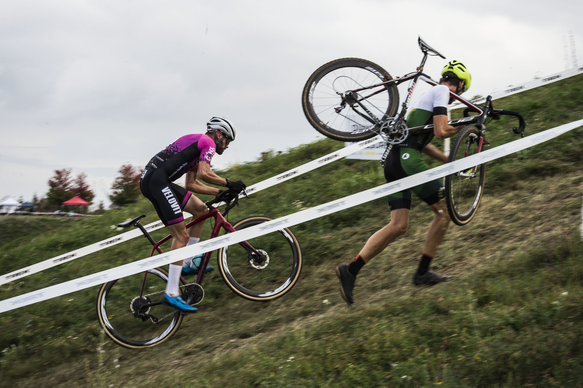 A cyclocross racer struggles to ride his bike up a steep hill, while another carries his bike.