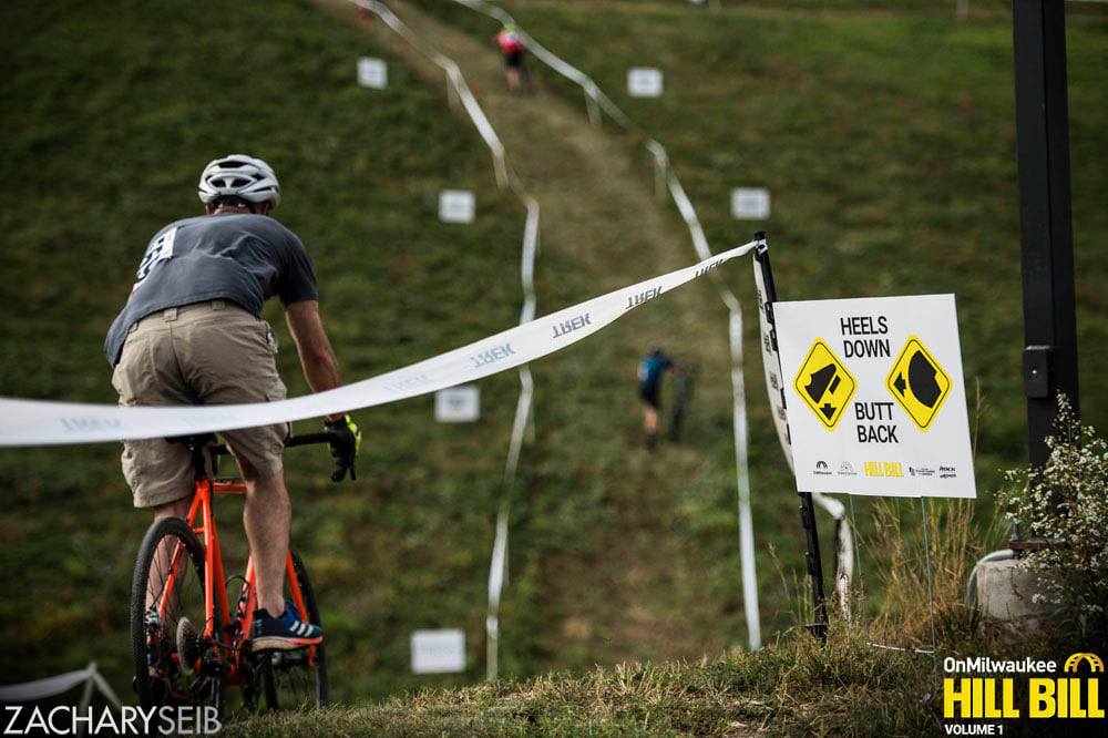 A cyclocross racer begins their descent down a steep hill next to a sign that reads 'Heels down, butt back.'