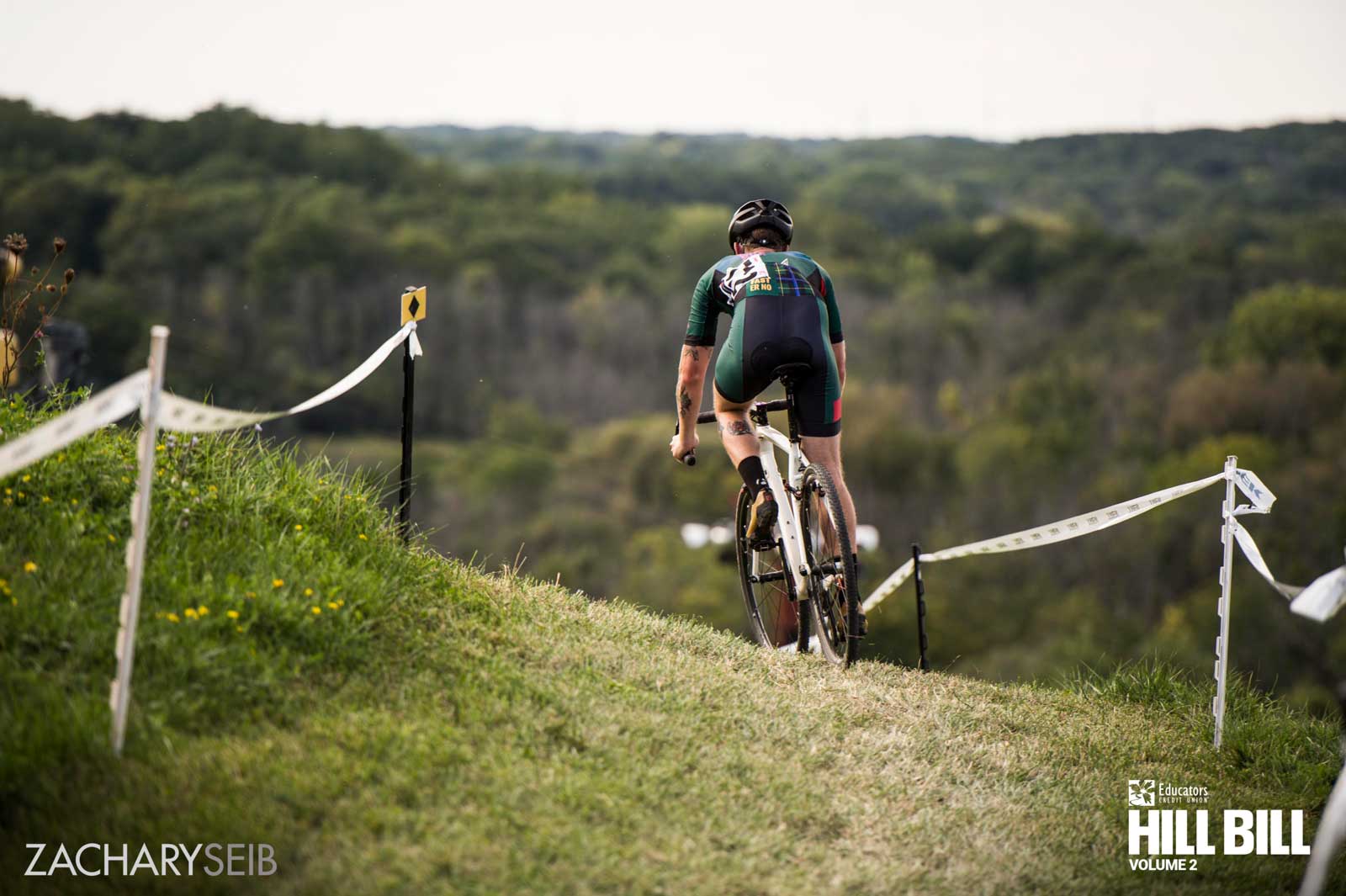 Behind a cyclocross racer about to drop down a descent overlooking a beautiful wooded area.
