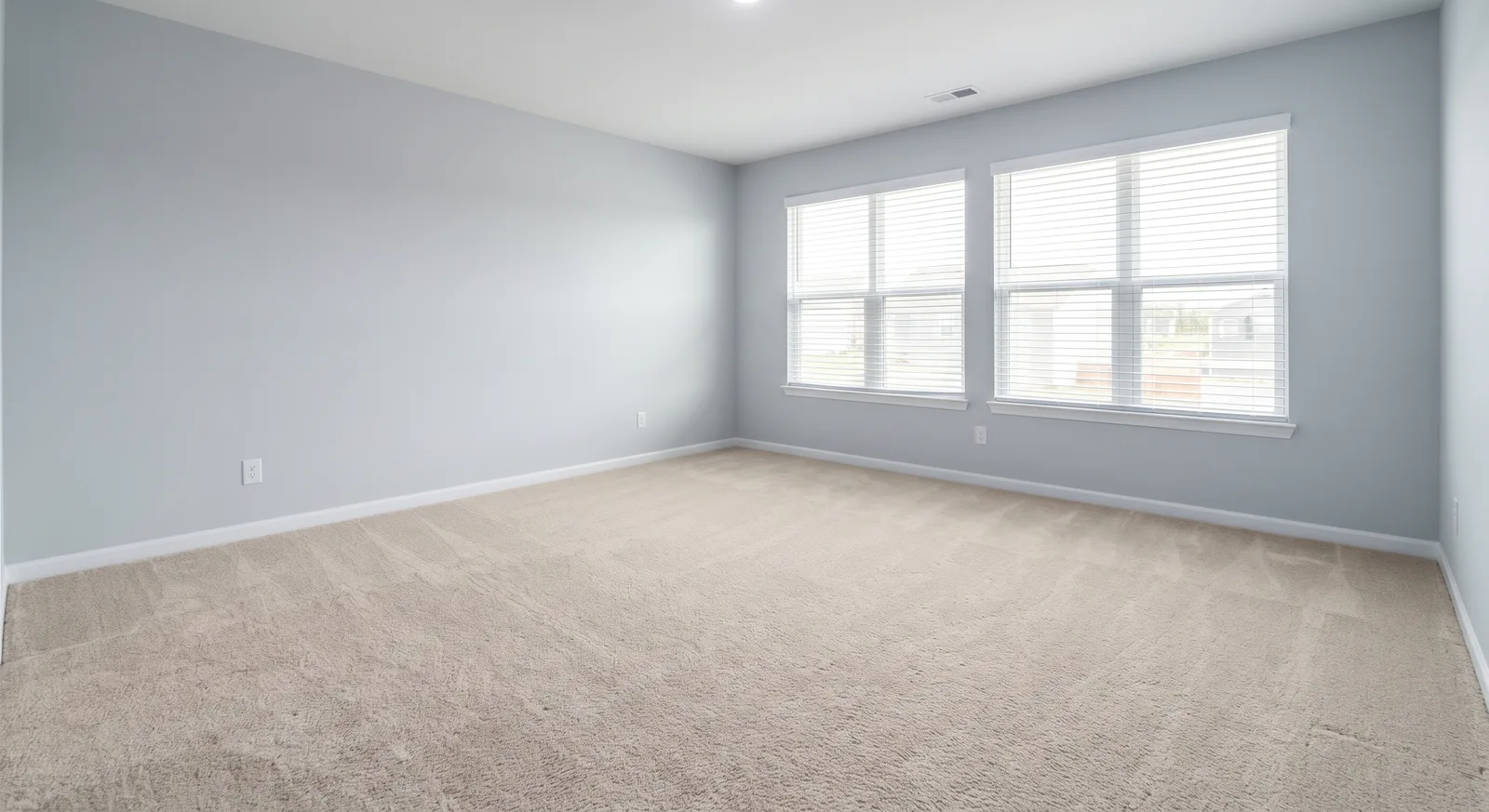 Before virtual staging: the same family room empty, with gray walls, beige carpet and two windows with white blinds.