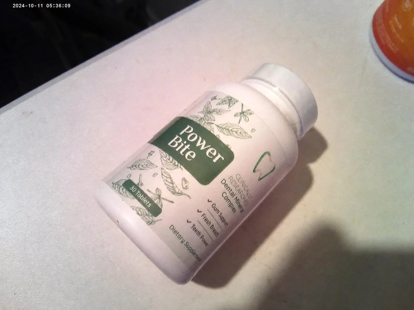 PowerBite bottle at an angle on a white table, green Power Bite label facing up, with a camera date-stamp in the corner