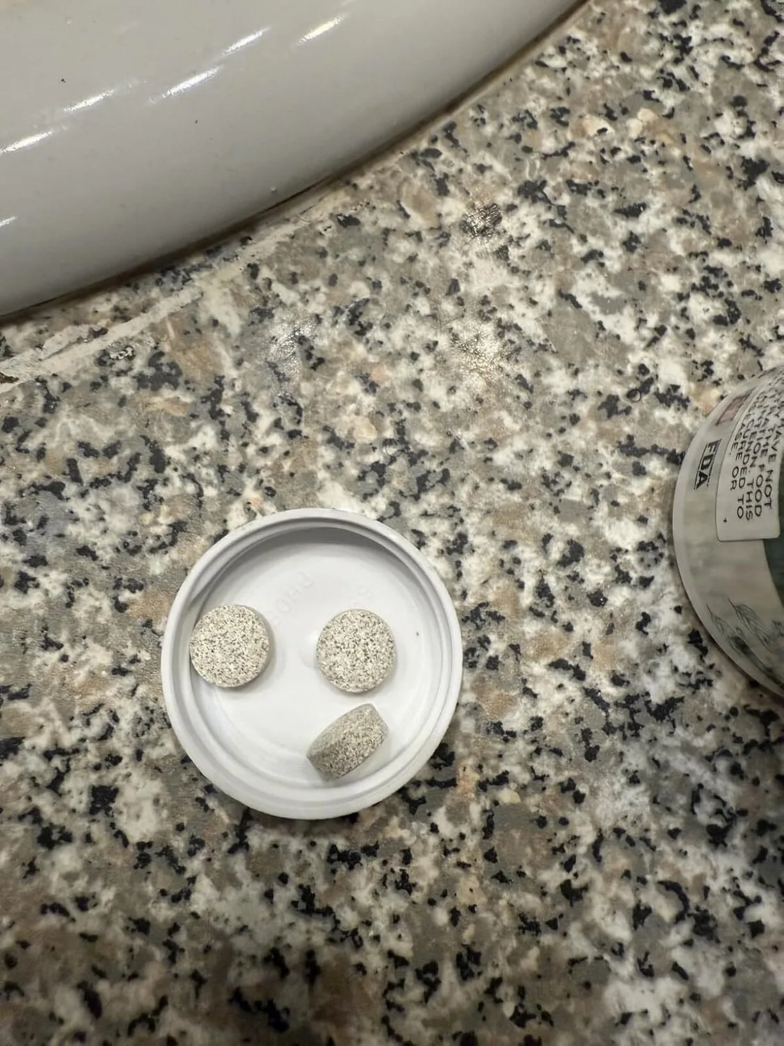 Three speckled tan PowerBite tablets in the white bottle cap on a granite counter, one on its edge showing thickness