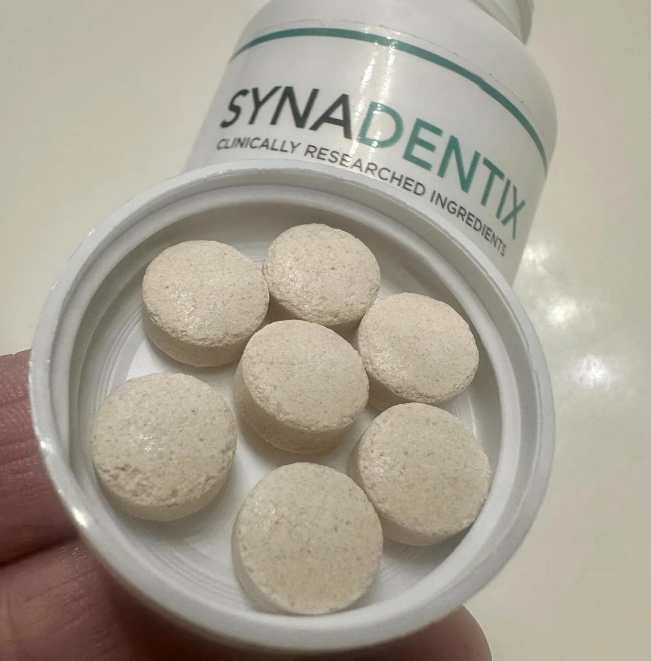 Seven off-white Synadentix tablets arranged on the white cap, with the tilted bottle and label behind them