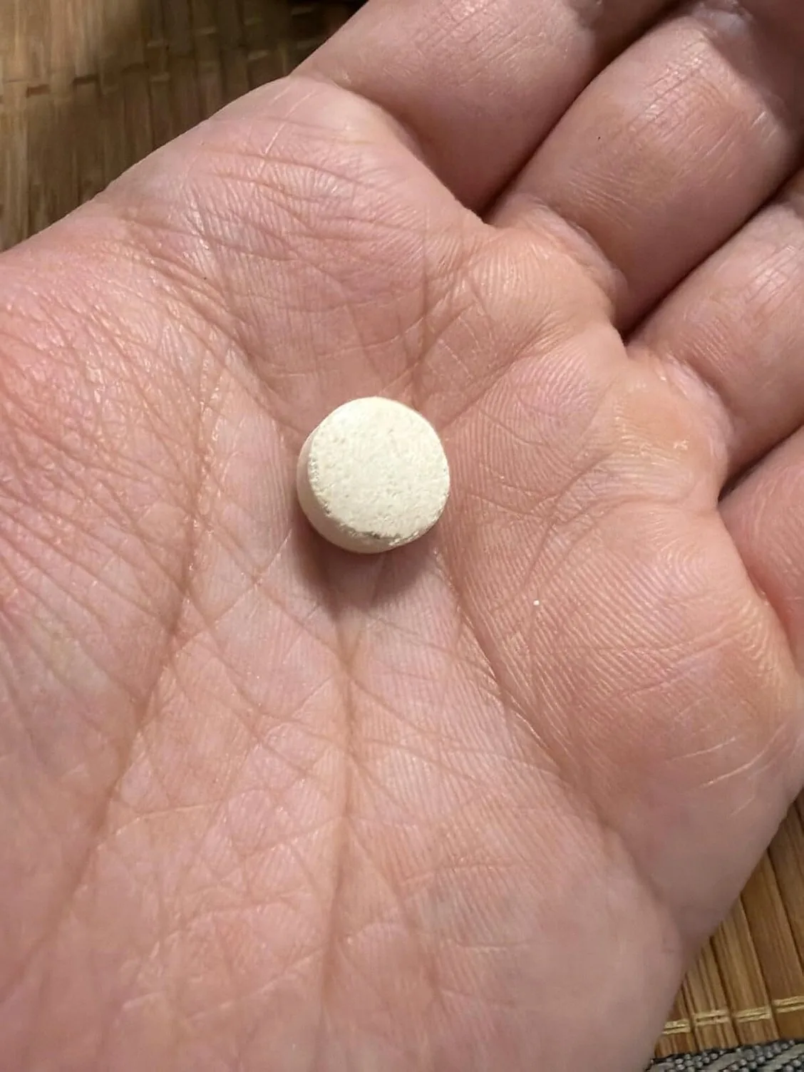 A single small off-white Synadentix tablet resting in an open palm over a bamboo mat, showing its round pressed shape
