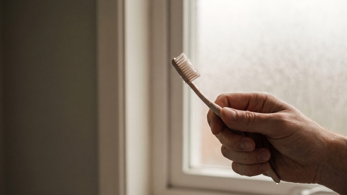 A hand holding a soft-bristle toothbrush by a bathroom window in morning light.