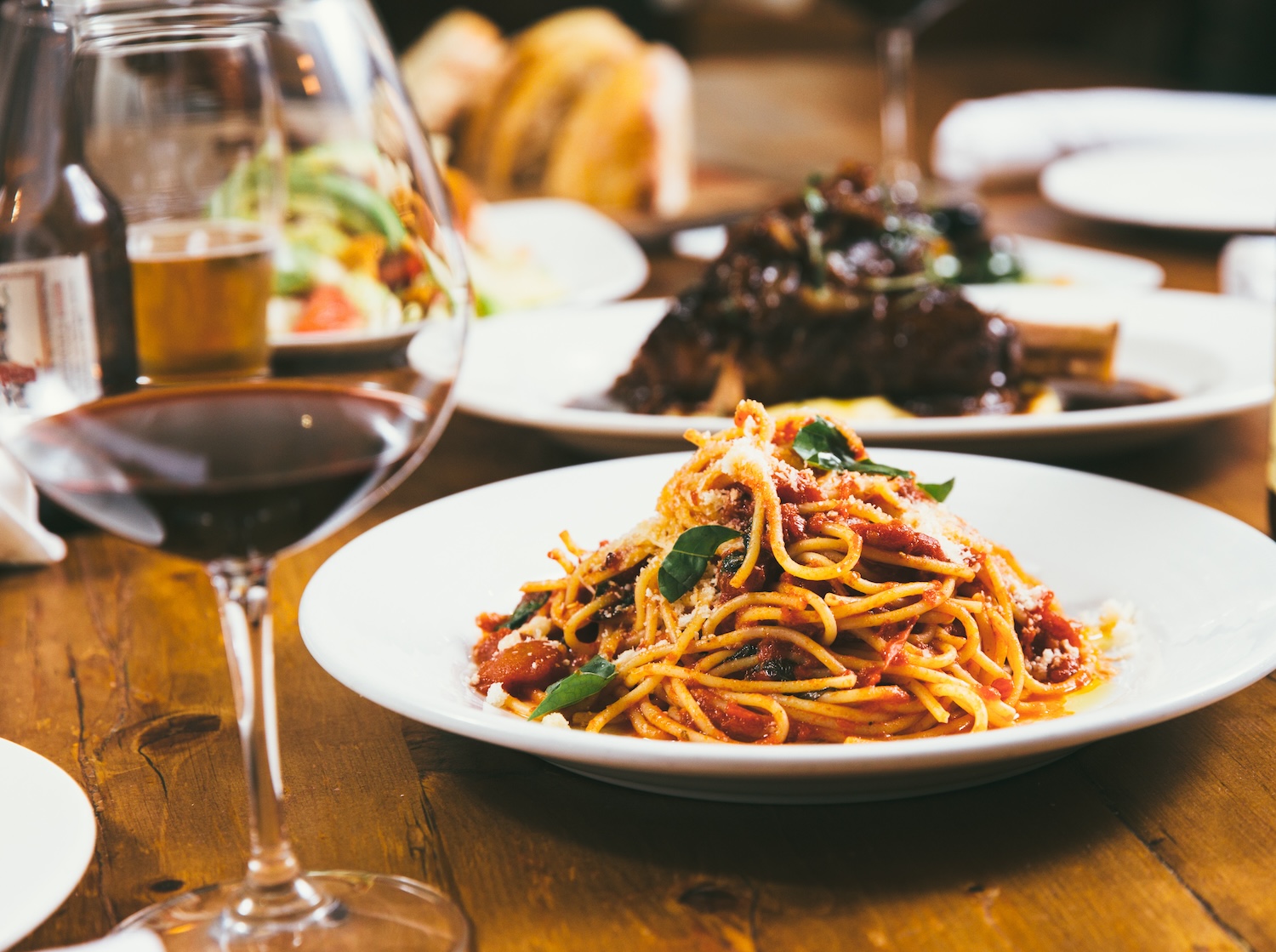 A plate of spaghetti pomodoro sits on a wooden farm tables surrounded by Italian dishes and red wine.