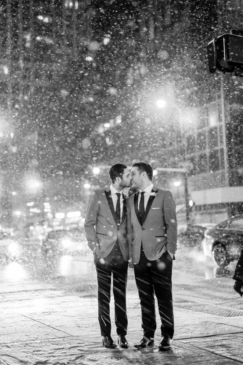 A just married couple share a kiss underneath falling snow outside Osteria Via Stato.