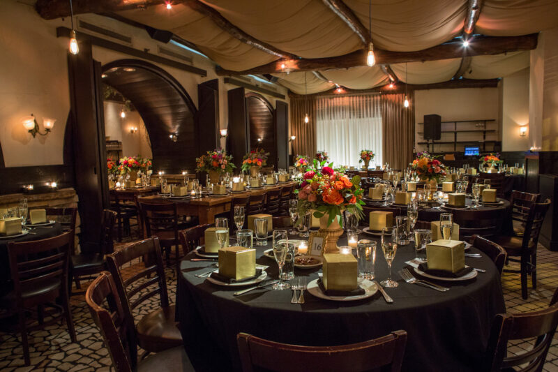 Round and long table in dining room set for wedding, with black linens, gold favor boxes and orange and red floral centerpieces