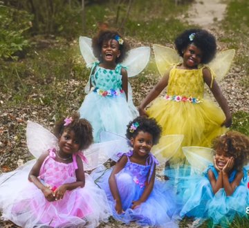 five young black girls are dressed as fairies in a lush green forest. the girl in the center is wearing a purple dress, while the girls on either side of her are wearing pink, blue, teal, and yellow dresses. they are all smiling and look like they are having fun.