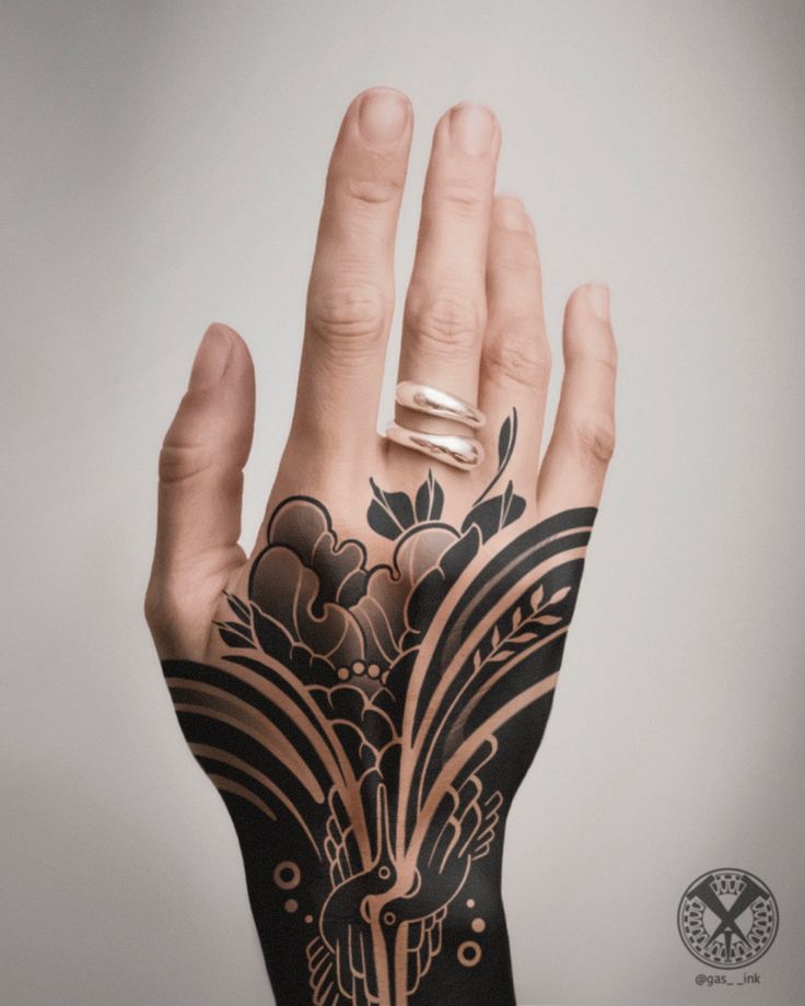 the image features a hand with a large black ink tattoo. the tattoo covers the back of the hand and extends onto the wrist. the design includes elements such as clouds, leaves, and birds. the hand has a silver ring on one of the fingers. the background is a plain white or light gray.