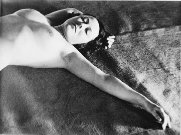 a black and white photograph of a nude woman lying on her side. she is positioned with her arm outstretched and her hand resting gently on the ground. her expression appears to be one of peaceful serenity. the image has a vintage and artistic quality, with visible grain and texture in the photograph. the lighting and shadow play create a sensual and intimate mood. the overall composition and aesthetic is reminiscent of classic fine art photography.
