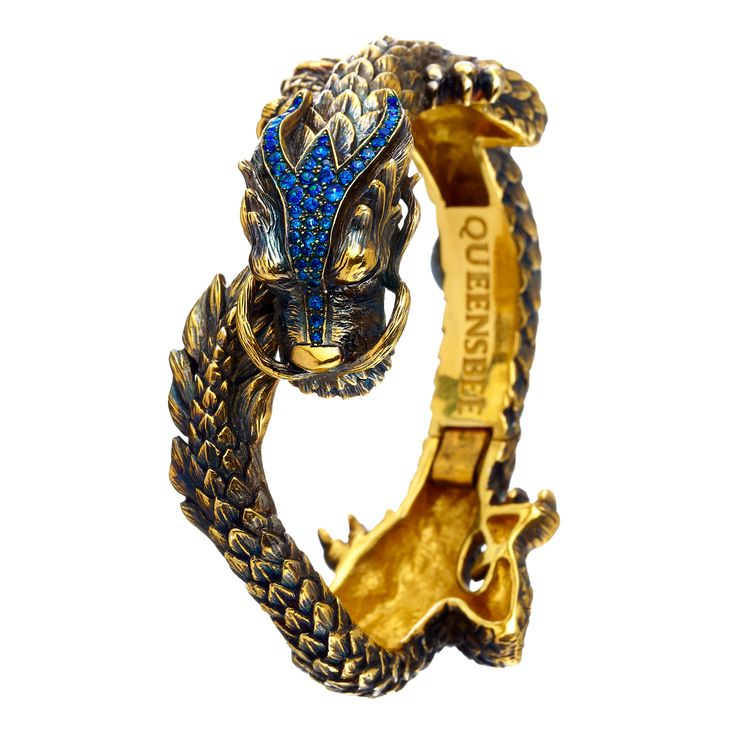 the image shows a gold dragon bracelet with blue crystals on its head. the bracelet is open, revealing the inside where the word "queensbee" is inscribed. the dragon's head is highly detailed with scales and sharp features.