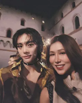 a closeup portrait of a young man and woman standing close together outdoors at night.  the man is wearing a stylish, darkcolored jacket with gold detailing, and the woman is wearing a lightcolored top. they are both smiling warmly at the camera. the background features a large, old stone building, possibly a castle or historical structure,  giving a romantic and somewhat mysterious ambiance to the image. the scene appears to be a nighttime event or celebration.