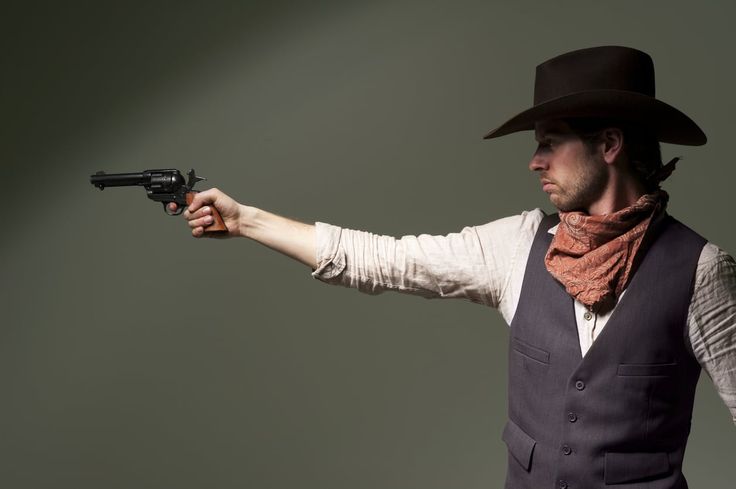 a man wearing a cowboy hat and vest holds a revolver in his right hand. he is looking away from the camera with a serious expression on his face. the background is a plain grey and green.