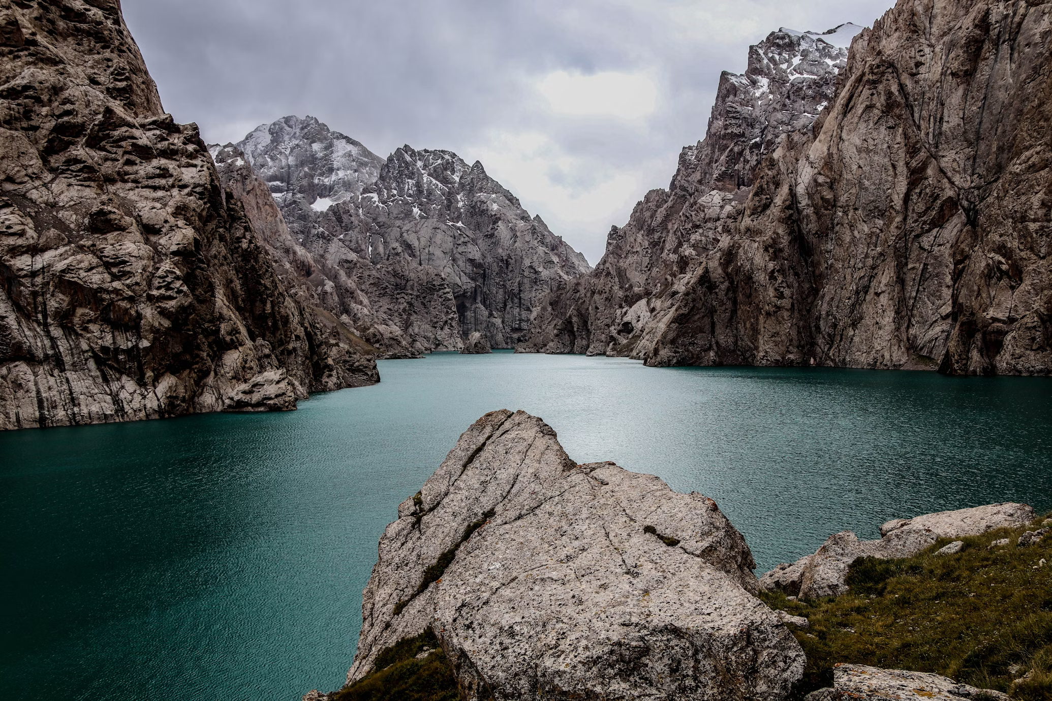 the image showcases a breathtaking mountain lake nestled between towering, rocky peaks. the lake's surface, a captivating turquoise hue, reflects the overcast sky, creating a serene and slightly moody atmosphere.  rugged, greybrown mountains, their slopes dusted with snow, frame the scene, emphasizing the grandeur of nature. a large, textured rock in the foreground provides a sense of depth and perspective.  the overall composition exudes a sense of tranquility and aweinspiring beauty.