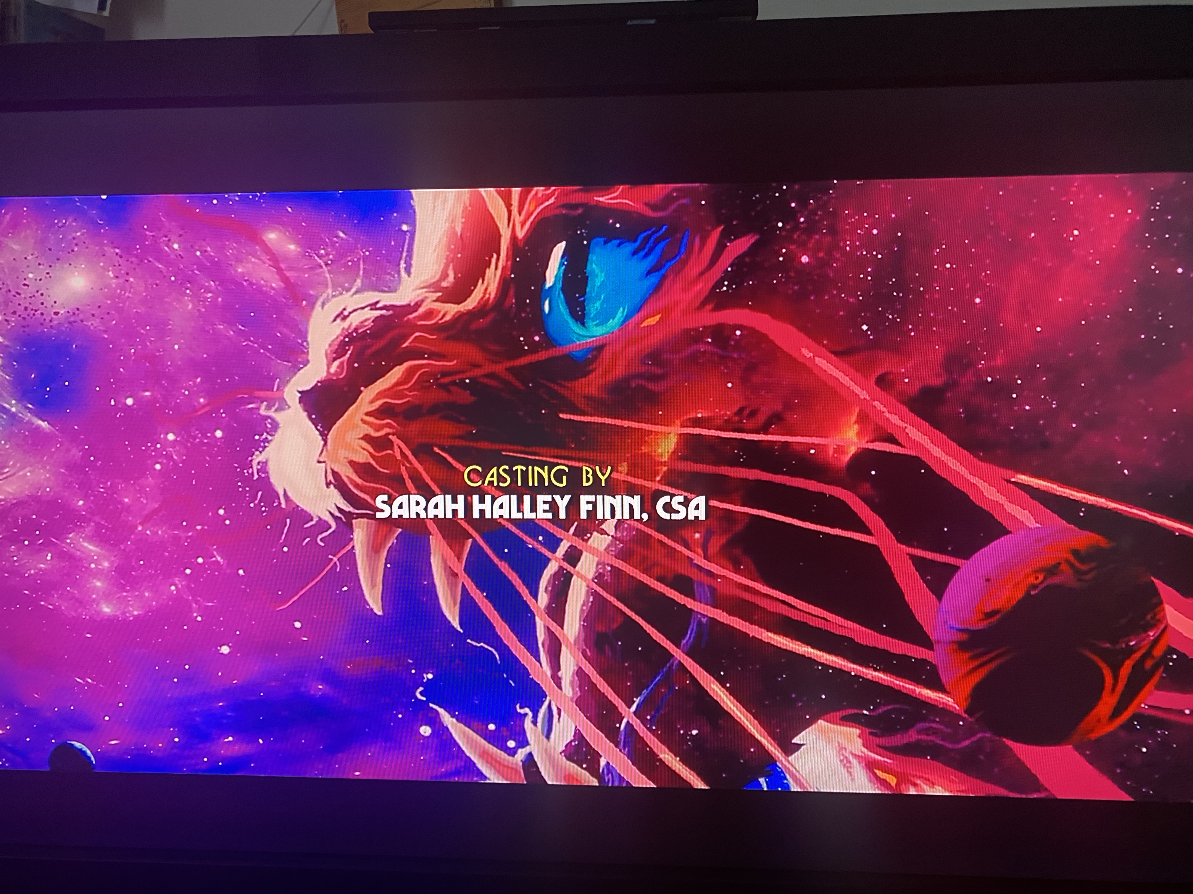 a still image of a movie's end credits. the background shows a space scene with a red and blue nebula filled with white stars. the foreground has an orange cat with a redtinged glow, with its eye visible, along with the mouth and whiskers. the text "casting by sarah halley finn, csa" is in white against a yellow background, indicating the casting director's name.