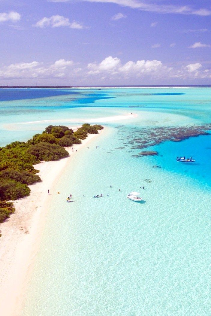 an aerial shot of a small, idyllic island surrounded by crystal clear turquoise water. the white sand beach is lined with lush green vegetation, and a few people are enjoying the water and sunshine. two boats are anchored in the shallows. the sky is a vibrant blue with fluffy white clouds.