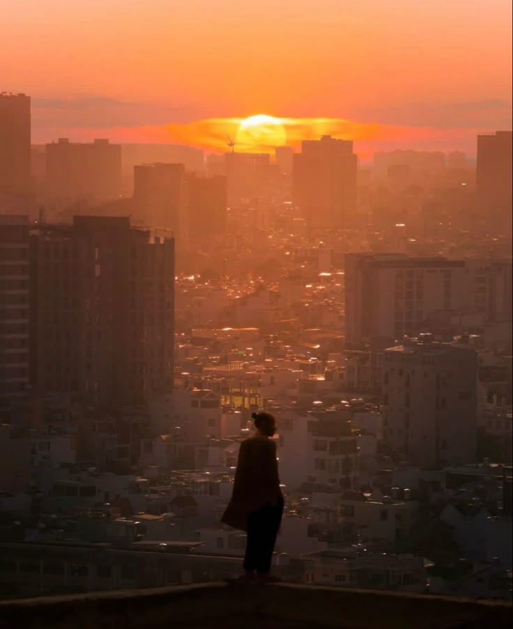 the image showcases a breathtaking sunset over a densely populated city. a lone female silhouette stands on a rooftop, seemingly contemplating the sprawling cityscape bathed in the warm, golden light of the setting sun. the city stretches out below, a vast expanse of buildings and structures fading into a hazy atmosphere. the overall mood is one of serene solitude and peaceful contemplation, heightened by the dramatic colors of the sky and the solitary figure against the urban backdrop. the sun, partially obscured by a cloud, is a striking feature in the center of the image.  the visual style suggests a slightly surreal or dreamlike quality.