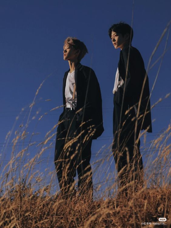 two young men wearing black suits with suspenders stand in a field of dry, golden grass. they are looking up towards the clear blue sky. the sun is shining brightly, creating a warm, golden glow and backlighting the figures. the image is taken from a low angle, emphasizing the vastness of the sky and the youthful energy of the subjects.