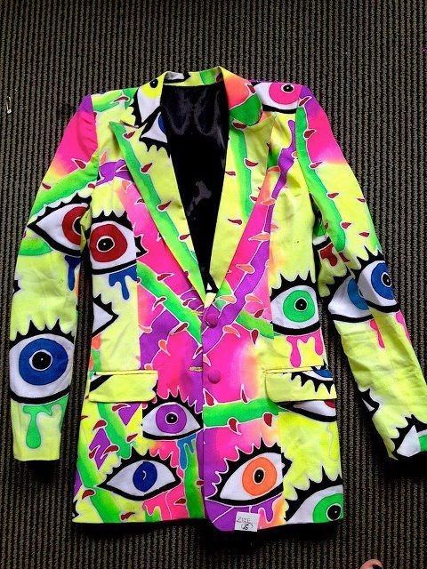 a brightly colored blazer jacket with an eye pattern is laid flat on a surface. the jacket is designed with neon shades of yellow, pink, purple, and green. the pattern features various stylized eyes with different colored irises. the jacket has a black lining, a doublebreasted closure with visible buttons, and two pockets.