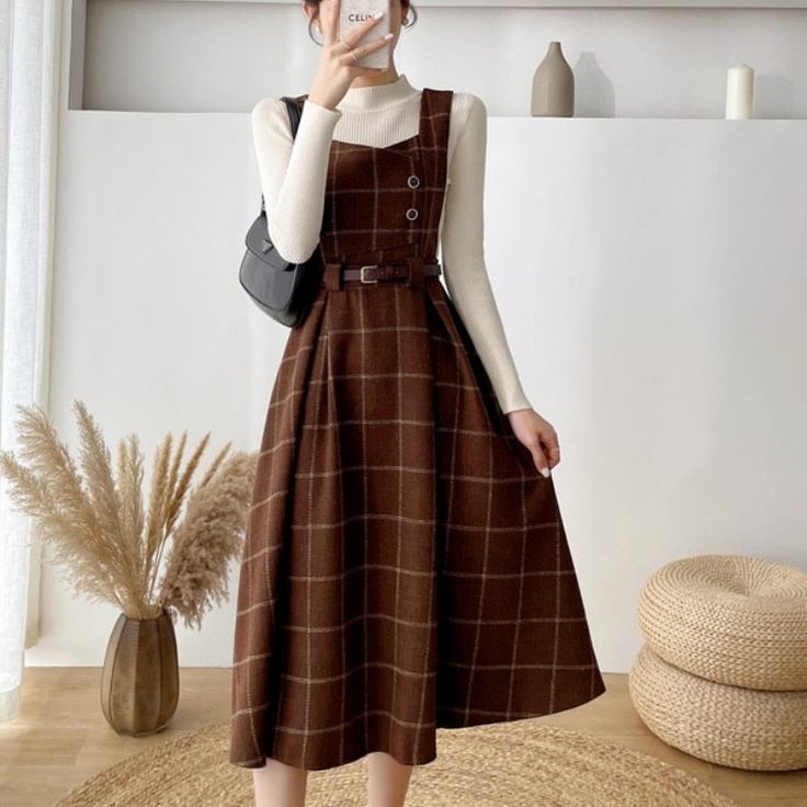 a woman is standing indoors, modeling a brown and white plaid dress. the dress has a square neckline, thick straps, a belt, and buttons down the front. she is also wearing a white longsleeve shirt underneath the dress. the dress falls below her knees, and her shoes are not visible in the photo. she is carrying a black purse and holding her phone up, as if taking a selfie. the background is a creamcolored wall with a framed picture hanging on it. there is a vase of dried flowers to the left of the woman and a woven stool to the right. the lighting is soft and natural.
