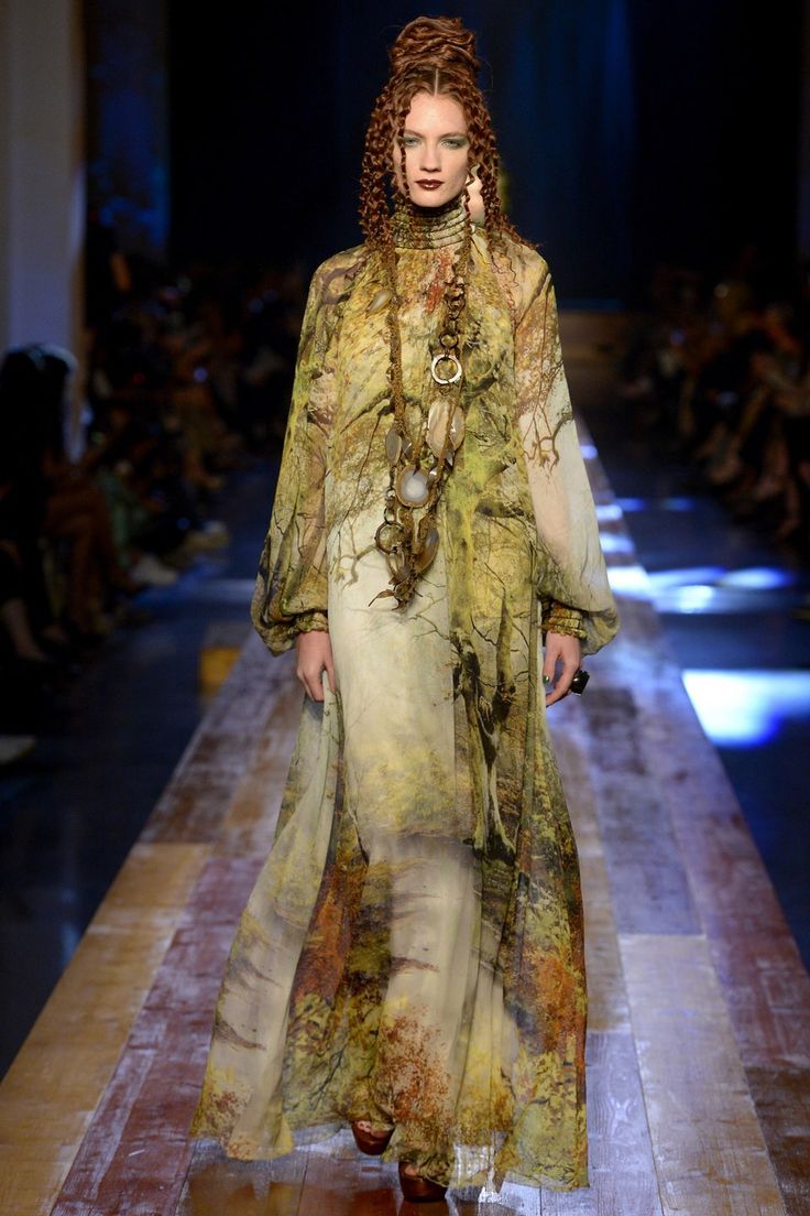 a female model with long, curled red hair is walking down a runway wearing a long, flowing dress with a floral pattern. the dress has long sleeves and appears to be made of silk.  she has a heavy necklace of woven beads and a belt around her waist. the background is blurred, but you can see audience members.