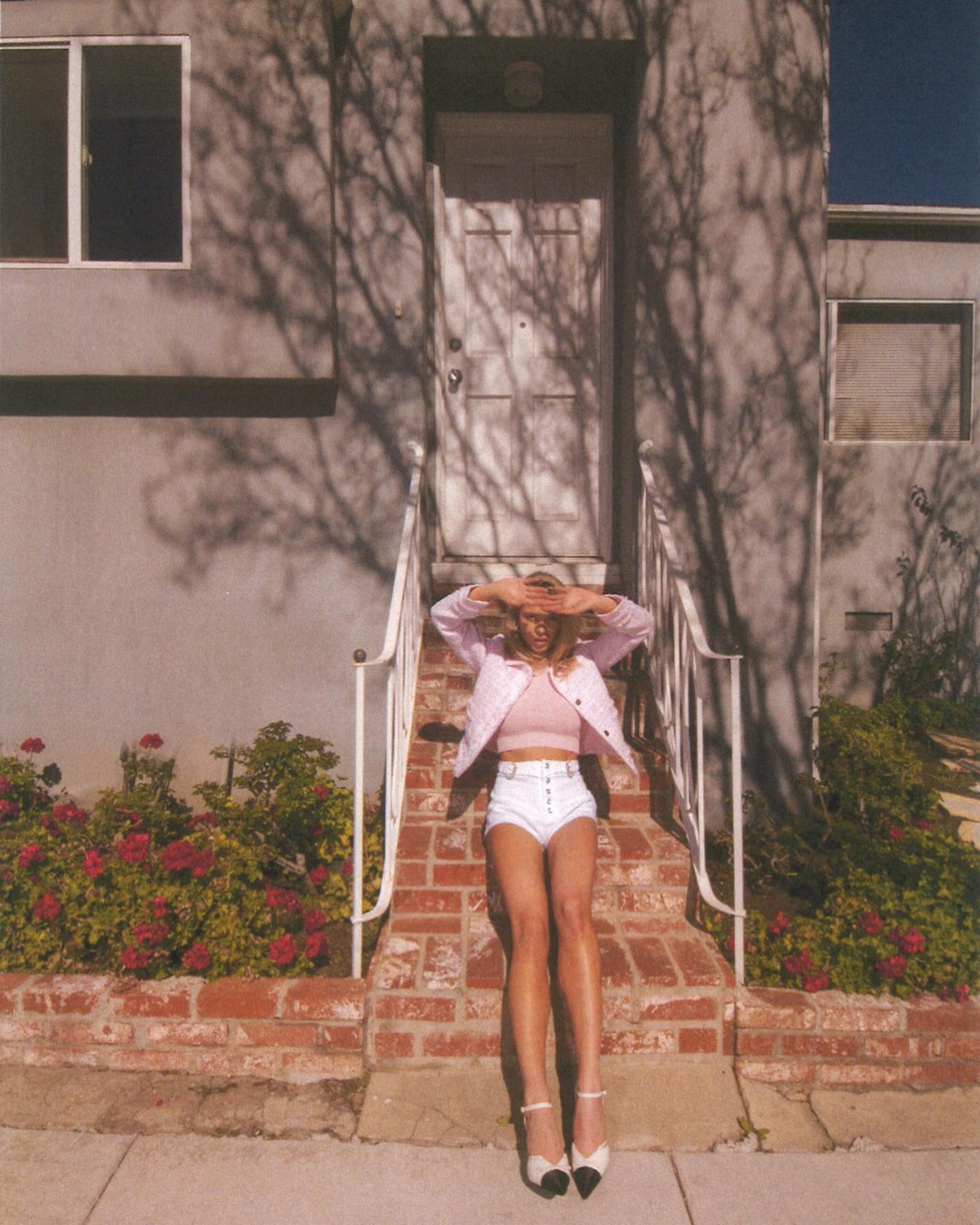 the image shows a young blonde woman sitting on brick steps in front of a house. she is wearing a pink jacket, white shorts, and white shoes. the style is reminiscent of the 1960s.  the sun is shining, casting shadows from a tree onto the building. the setting appears to be a residential area, possibly in california, given the architecture and landscaping. the overall mood is casual and summery.  the woman is posing, with her hands shielding her eyes from the sun.  the image is bright and has a slightly vintage or filmlike quality.