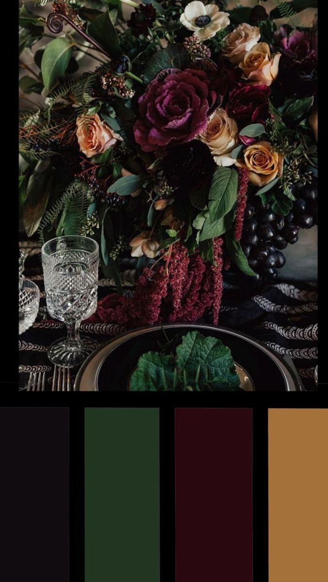 a moody and elegant wedding centerpiece with dark green, maroon, and brown accents. the centerpiece is made up of dark green and burgundy flowers and grapes, set on a table with black and white linens. there are two clear glass goblets on the table as well as silverware. the dark colors and rich textures create a romantic and spooky atmosphere.