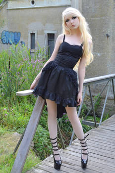 a young blonde woman in a black gothic lolita dress with lace details and ruffles poses on a wooden bridge outdoors. she is wearing highheeled shoes and her pale skin contrasts with her dark clothing.  the background features overgrown vegetation and an old stone building with graffiti.