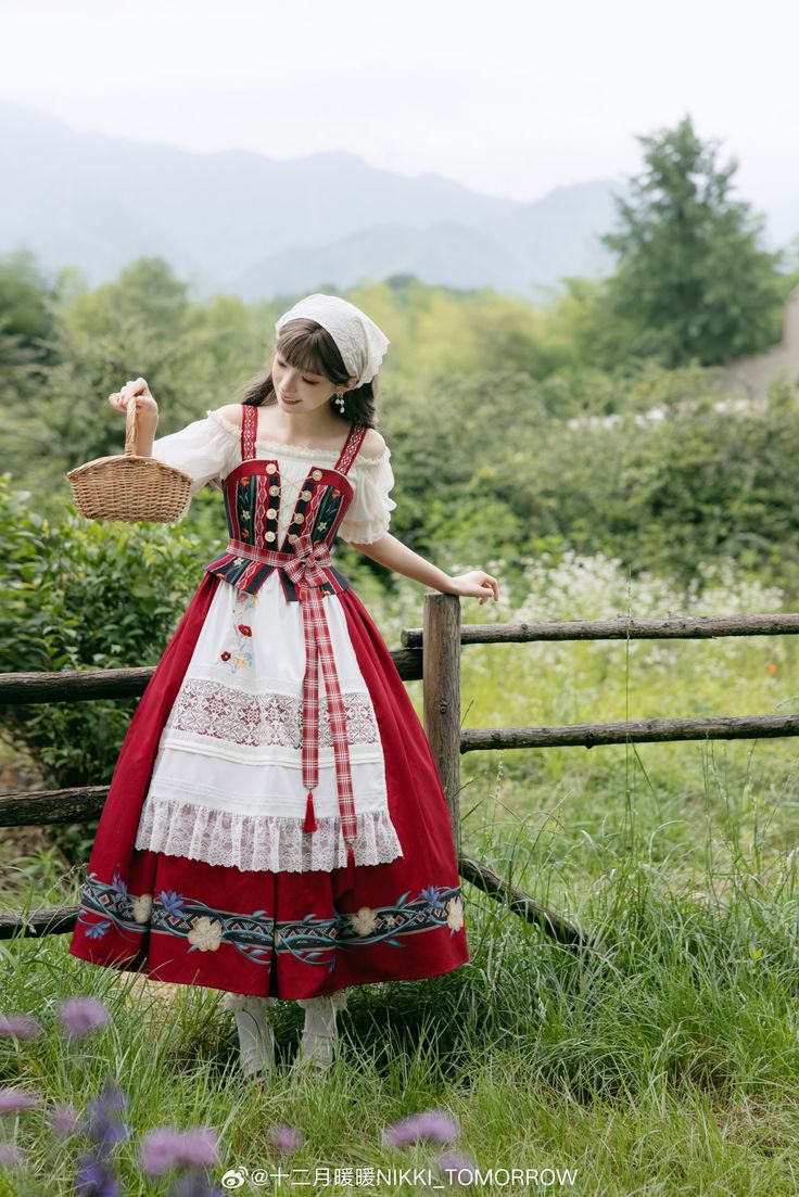 a young woman in a red and white lolita fashion dress stands in a field, holding a basket. she is leaning against a wooden fence, and there are mountains in the background. the lighting is natural and overcast, and the perspective is eye level.