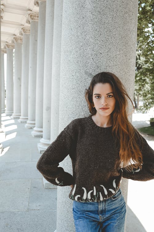 a portrait of a woman standing next to a large stone column. she's wearing a dark brown knit sweater with a white floral pattern around the bottom hem and cuffs, along with light blue jeans. her long brown hair is styled in loose waves, and she looks directly at the camera. in the background, there's a row of similar stone columns and some greenery, suggesting an outdoor setting near a building with classical architecture.
