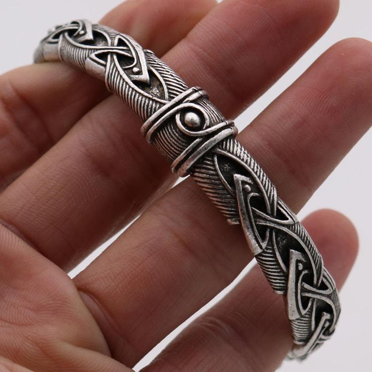 a closeup shot of a hand holding a silver bracelet with intricate celtic knot designs. the bracelet is made of metal and features a detailed pattern with a prominent knot motif. the hand is slightly blurred in the background, focusing attention on the bracelet's design and texture.