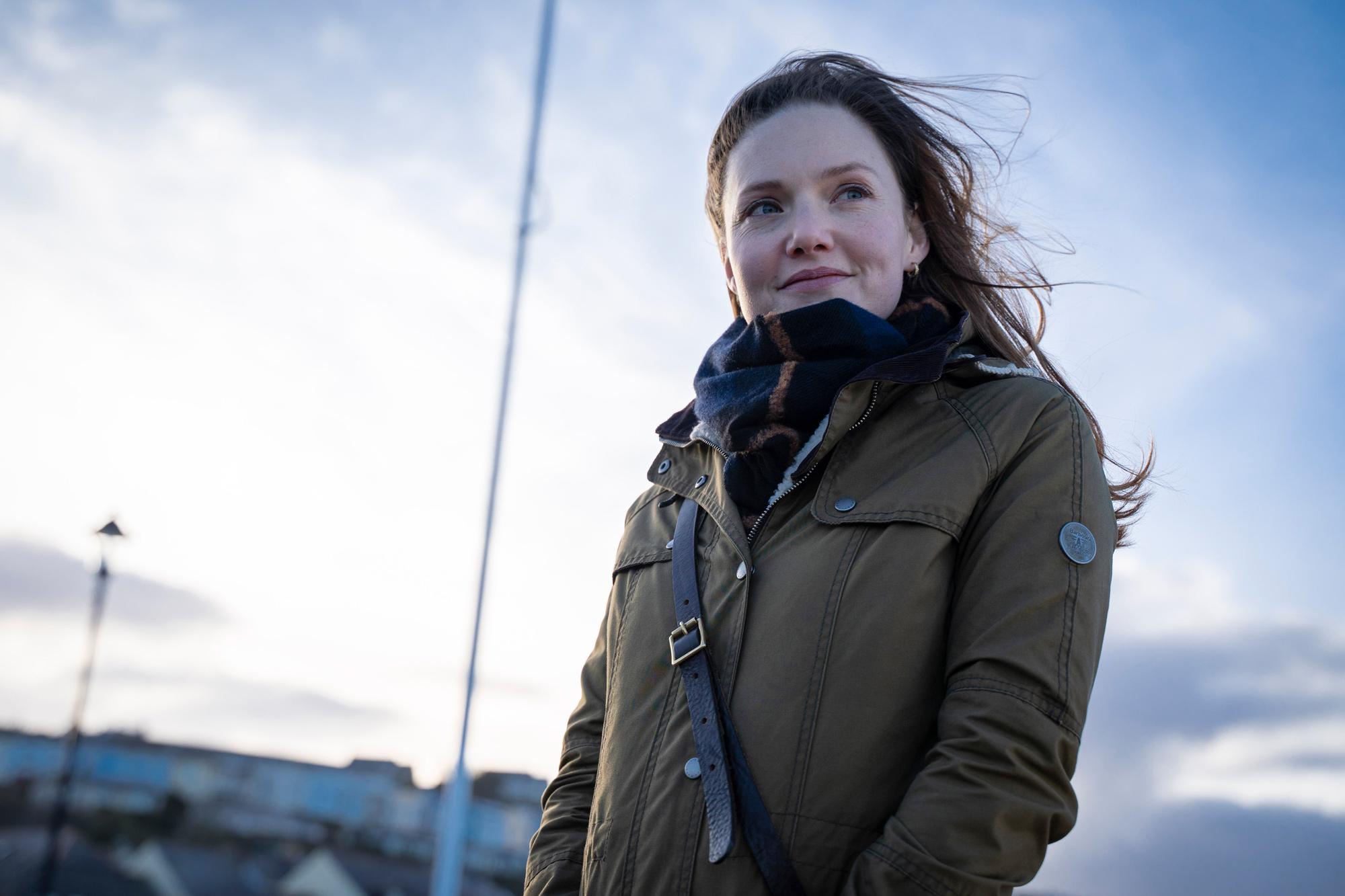 a medium shot of a woman standing outdoors on a somewhat windy day. her hair is blowing in the wind. she's wearing an olive green jacket and a dark patterned scarf.  she's looking off to the side and appears happy. the background is blurred but shows a city skyline and cloudy sky.