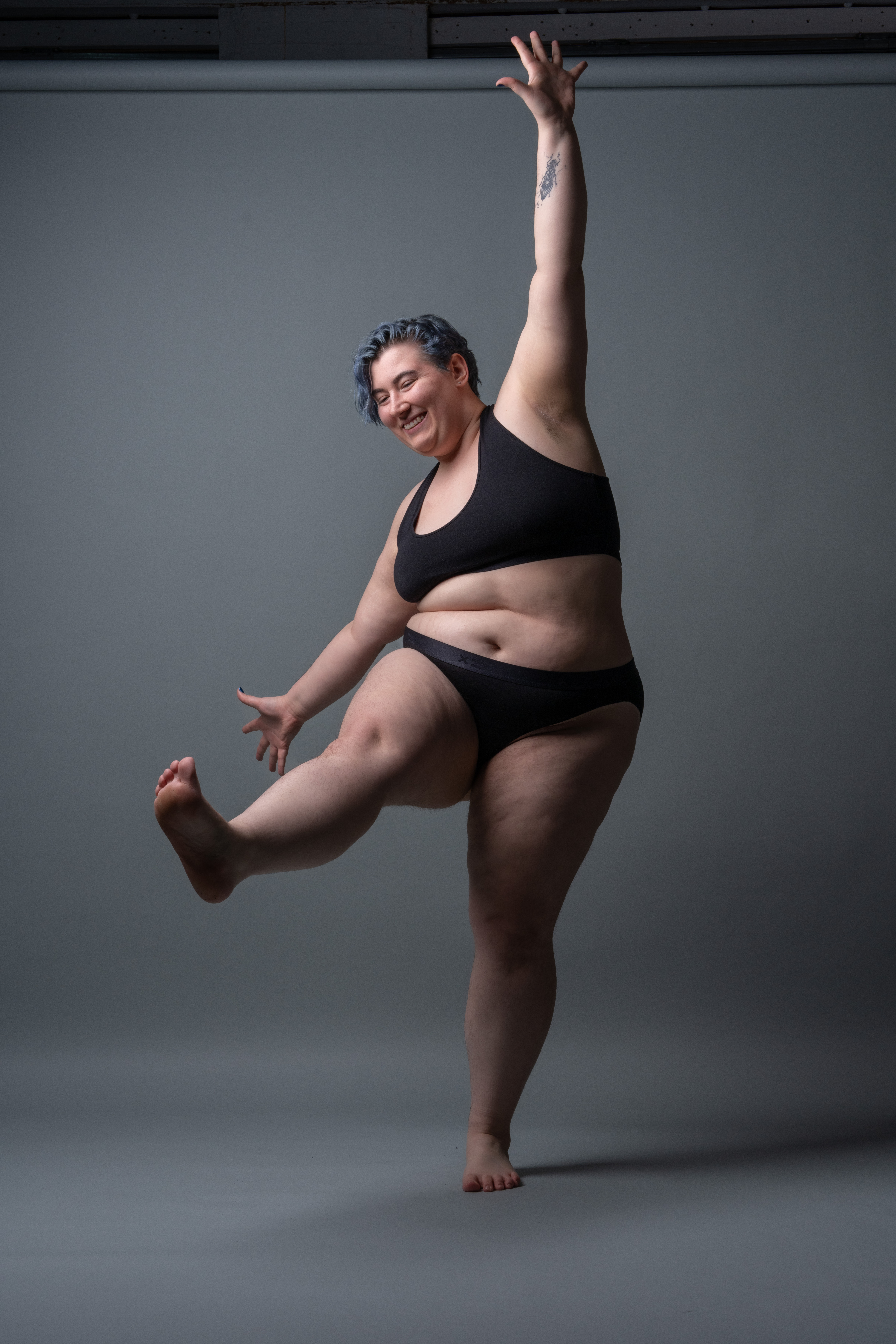 an image shows a smiling plussize woman with blue hair, wearing a black sports bra and underwear, barefoot and dancing against a gray backdrop. she has one arm raised and a leg lifted. the woman has a tattoo on her raised arm. she is expressing joy and body positivity.