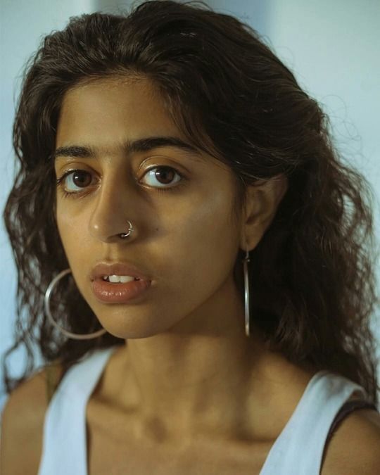 a closeup portrait of a young woman with long, dark, curly hair.  she has brown skin and is wearing simple silver hoop earrings and a small nose ring. her expression is serious and she is looking directly at the camera. the background is a plain, light color, drawing focus to the woman's face and features. she is wearing a simple offwhite tank top. the overall impression is one of natural beauty and a candid portrait.