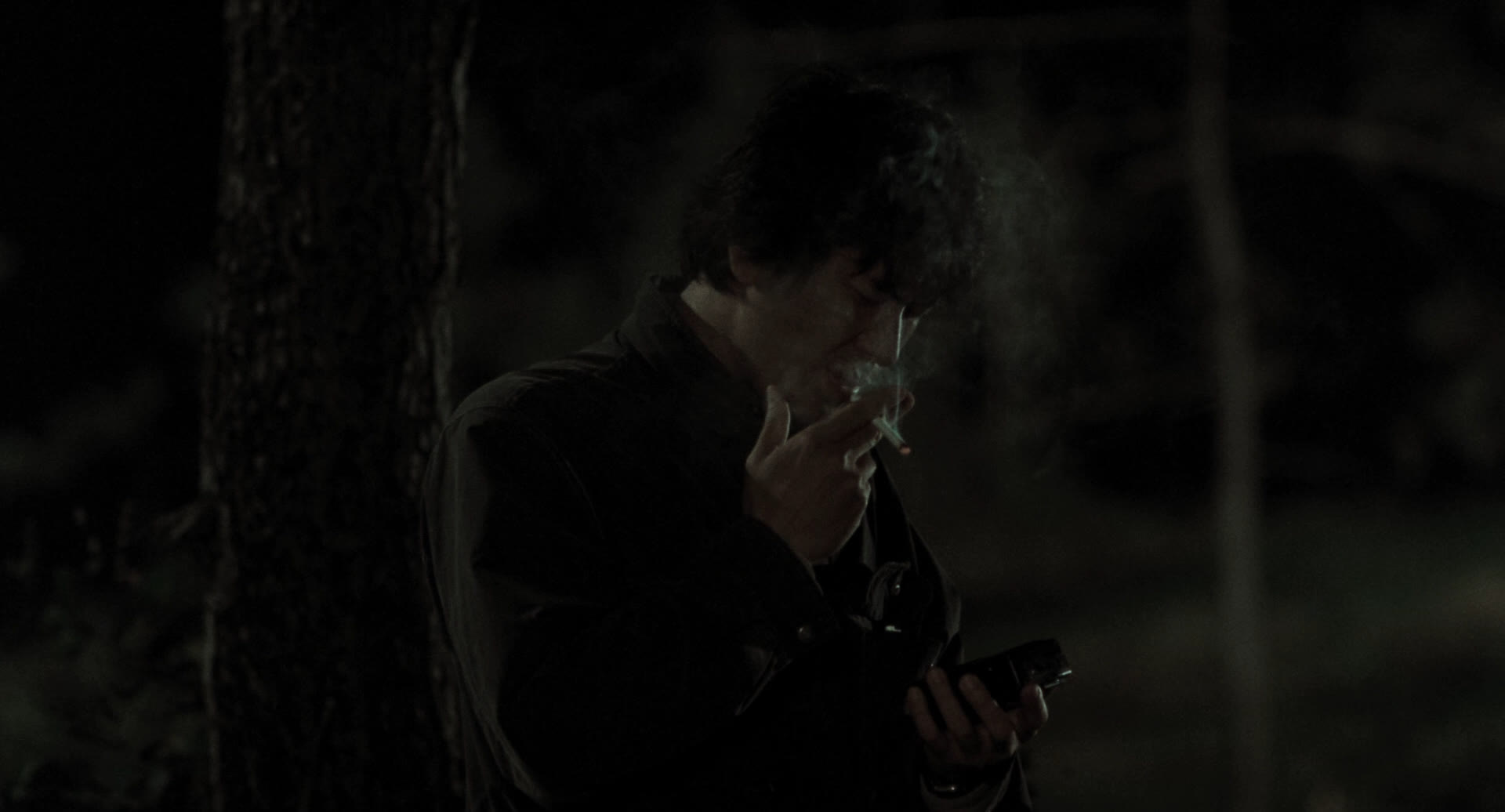 a man with dark, curly hair stands smoking a cigarette in a dimly lit forest at night. he's wearing a dark jacket and is holding something small and rectangular in his left hand. the overall atmosphere is mysterious and moody.