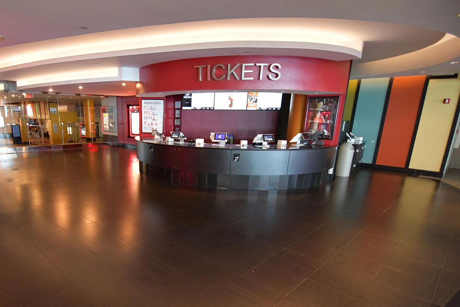 a photo of a mostly empty movie theater lobby. the image shows the ticket booth, a concession stand in the background to the left and a colorful striped wall on the right. the floor is dark brown tile and reflects the light from the ceiling.