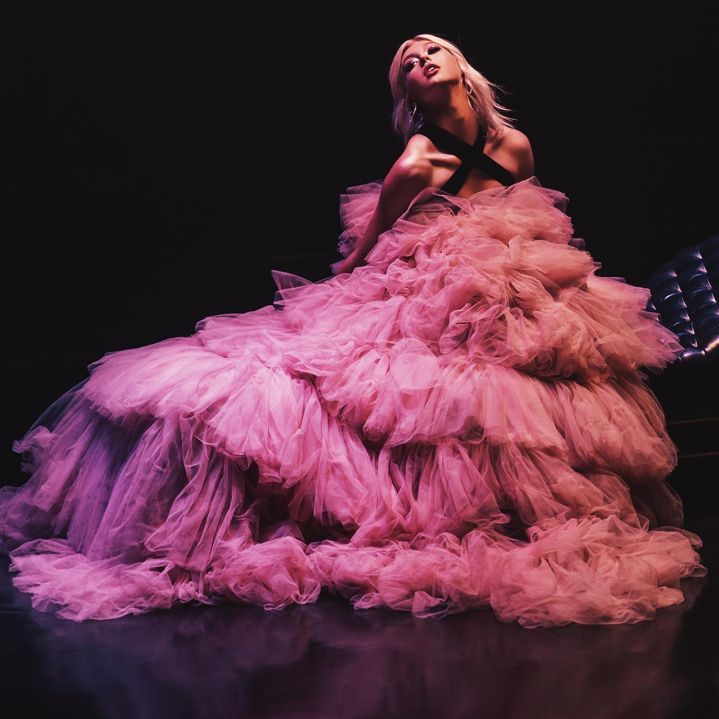 a woman with blonde hair is posing in a voluminous pink tulle gown against a dark background. she is looking upwards, creating an elegant and glamorous portrait. the dress features layers of ruffles.