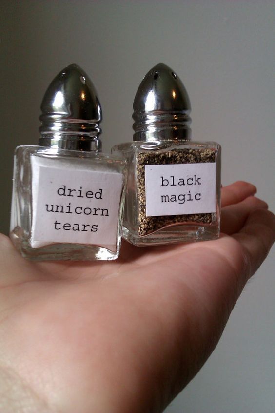 the image shows two clear glass salt and pepper shakers with metal tops held in someone's hand. the shaker on the left is labeled "dried unicorn tears" and contains a white powder. the shaker on the right is labeled "black magic" and contains a dark, granular substance. the image is a humorous take on common kitchen condiments.