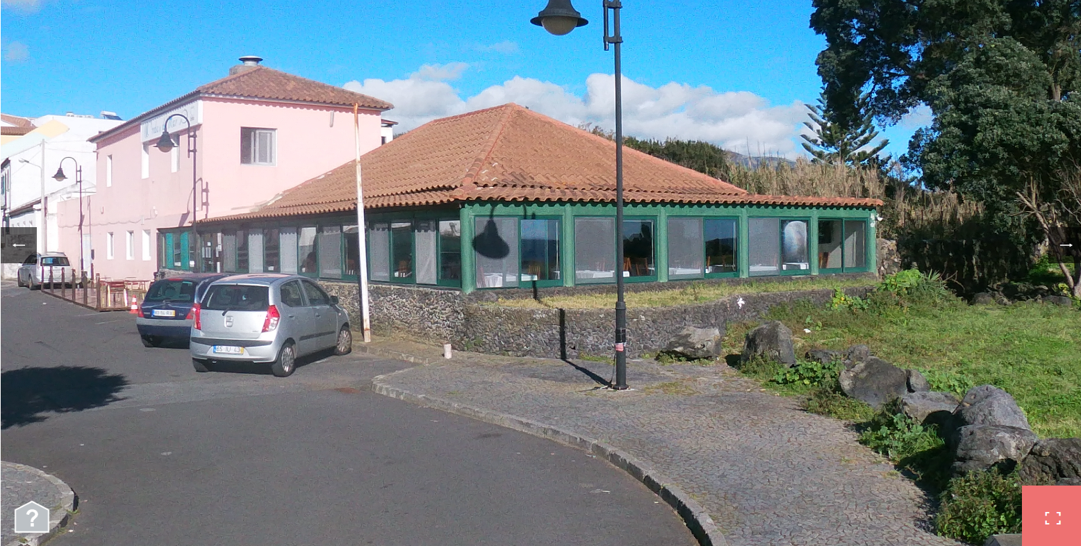 a view of a cafe or restaurant building on the left side of the image. the cafe has a brown tiled roof and a wall that is a pale pink color.  the building's windows are large and have a light green trim. there are small,  round windows below the large ones.  the cafe has a large,  partially covered patio with a stone wall  and a metal roof. the cafe is located along a paved street,  and  there are a couple of cars parked in front of it. there is a lamppost near the cafe,  and  a small sign at the front of the cafe.  there are trees to the right side of the image,  and  there is green foliage and grass around the cafe.  the sky is blue,  and  there are clouds in it. the day is sunny.