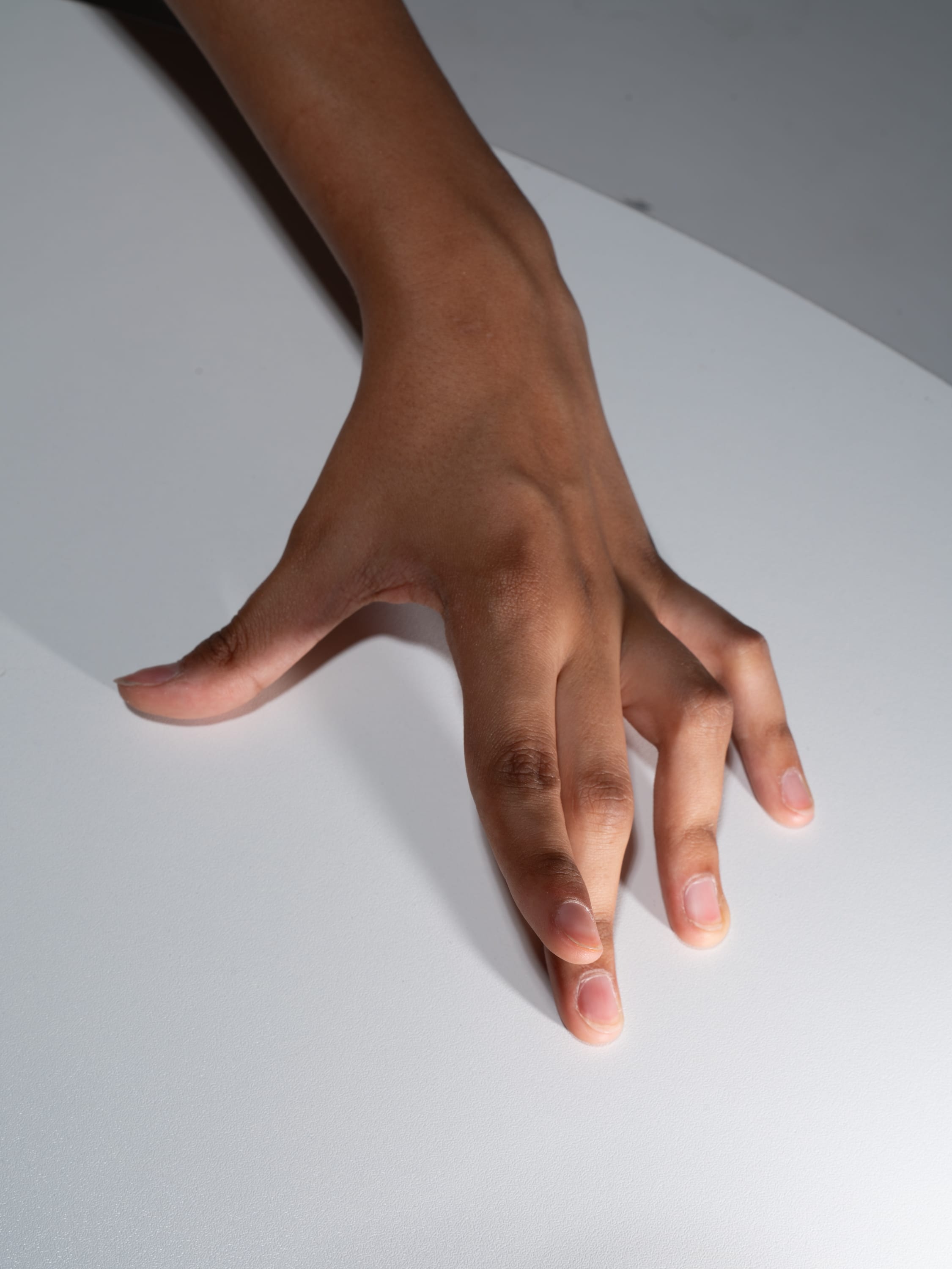 a hand with dark skin is extended on a white surface, with fingers slightly curled inwards. the light source appears to be from above, casting subtle shadows on the hand and surface. the image focuses on the hand and its details, capturing the texture of the skin and the position of the fingers.