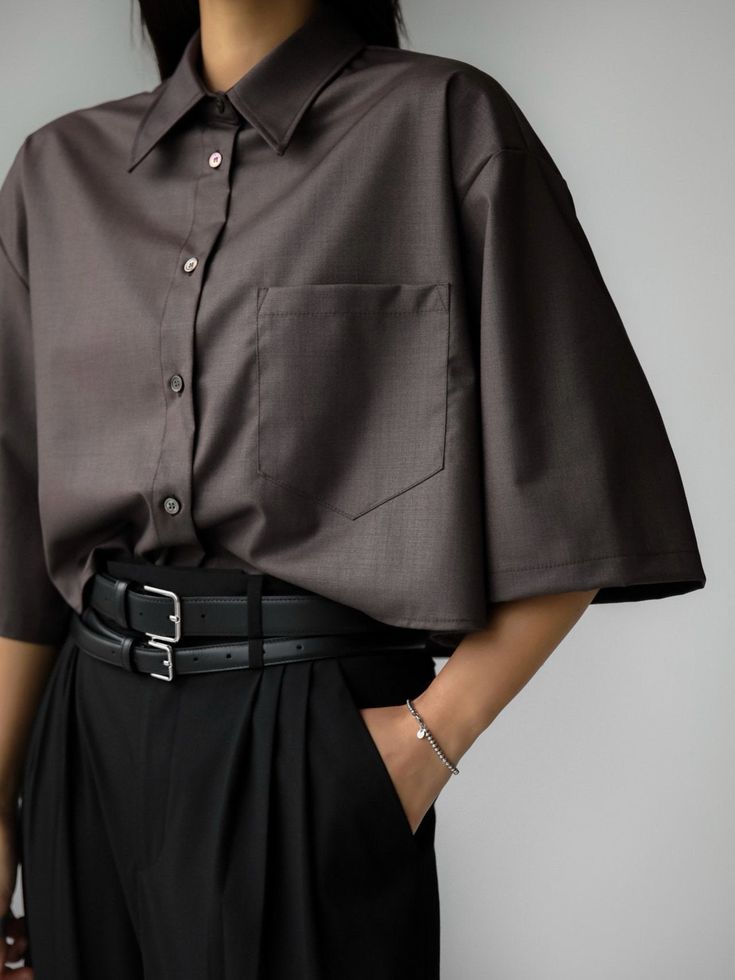 a woman is wearing a brown, oversized buttondown shirt with black pants and a black doublebuckle belt. she has a silver bracelet on her wrist and her hand is in her pocket. she is standing in front of a white background.