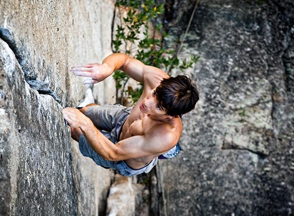 a man is rock climbing on a steep, rocky cliff face. he is wearing shorts and has chalk on his hands, showing his determination to reach the top. the rock face is textured and gray, with hints of green vegetation in the background. the man appears focused on the challenge before him.