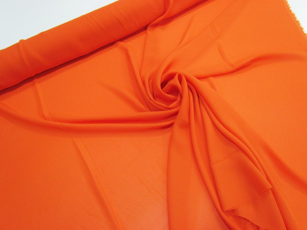 the image shows a roll of smooth, orange fabric draped on a white surface. the fabric is likely a lightweight material. the lighting appears to be indoor and artificial.