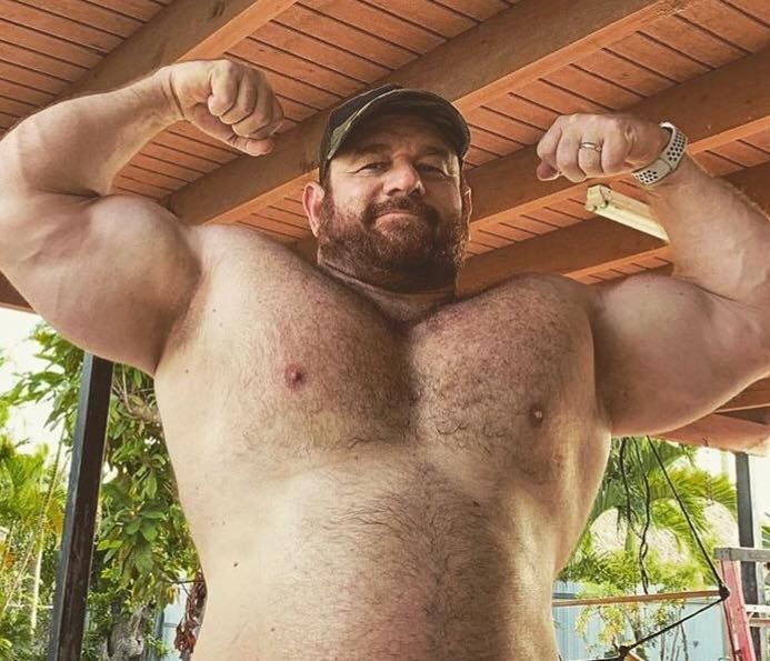 a muscular man with a beard is flexing his biceps while wearing a hat and a watch. he has a hairy chest and is standing outdoors.